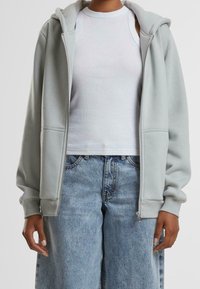 Personne portant un sweat à capuche gris clair zippé sur un haut blanc sans manches et un jean bleu taille haute, ample, debout avec les mains détendues.
