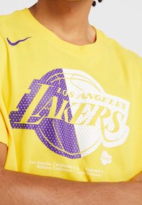 Żółta bawełniana koszulka z fioletowo-białym logo Los Angeles Lakers, grafiką koszykówki i szczegółami tekstowymi. Projekt z okrągłym dekoltem.