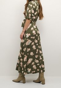 Robe florale longue vert olive avec de courtes manches bouffantes, mettant en avant des motifs de fleurs roses et bleues, et un détail de taille nouée. Associée à des bottes à talons beiges.