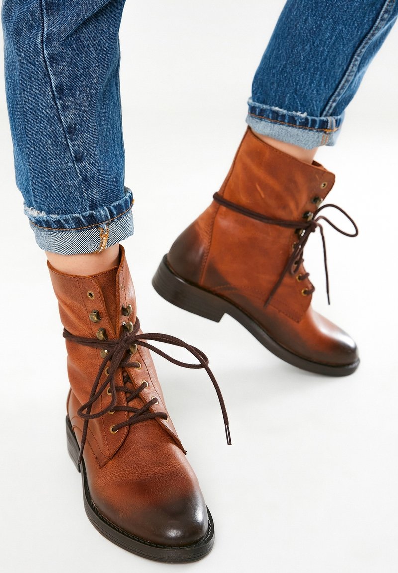 Bottes en cuir marron à lacets portées avec un jean bleu retroussé sur fond blanc.