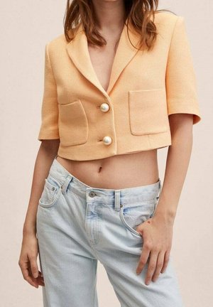 Femme portant un blazer court texturé couleur pêche avec des boutons en perles et un jean taille haute bleu clair, main dans la poche.