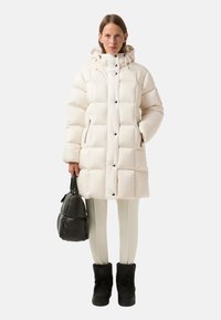 Puffy white coat with a hood, featuring large quilted sections, zippered side pockets, and black snap buttons; paired with black boots and a bag.