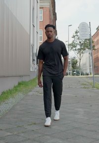 Svart t-shirt med tryckt grafisk motiv, svarta byxor och vita sneakers. Går på en asfalterad trottoar med byggnader i bakgrunden.