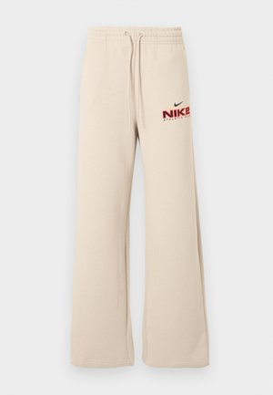 Röyhkeät beiget collegehousut, joissa on elastinen vyötärönauha ja kiristysnauhat. Vasemmassa reisessä on punainen ja musta Nike Athletic Club -logo.