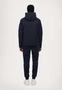 Giacca a vento blu navy con una texture liscia, abbinata a pantaloni jogger coordinati. Sneakers bianche alla base, viste da dietro.