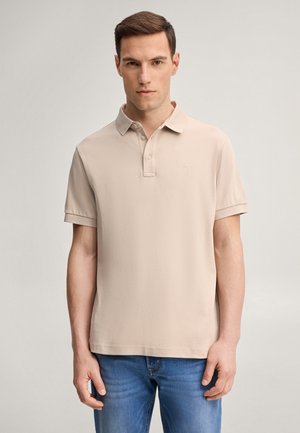 Man met kort bruin haar draagt een beige poloshirt met korte mouwen en blauwe jeans, staand tegen een effen lichte achtergrond.