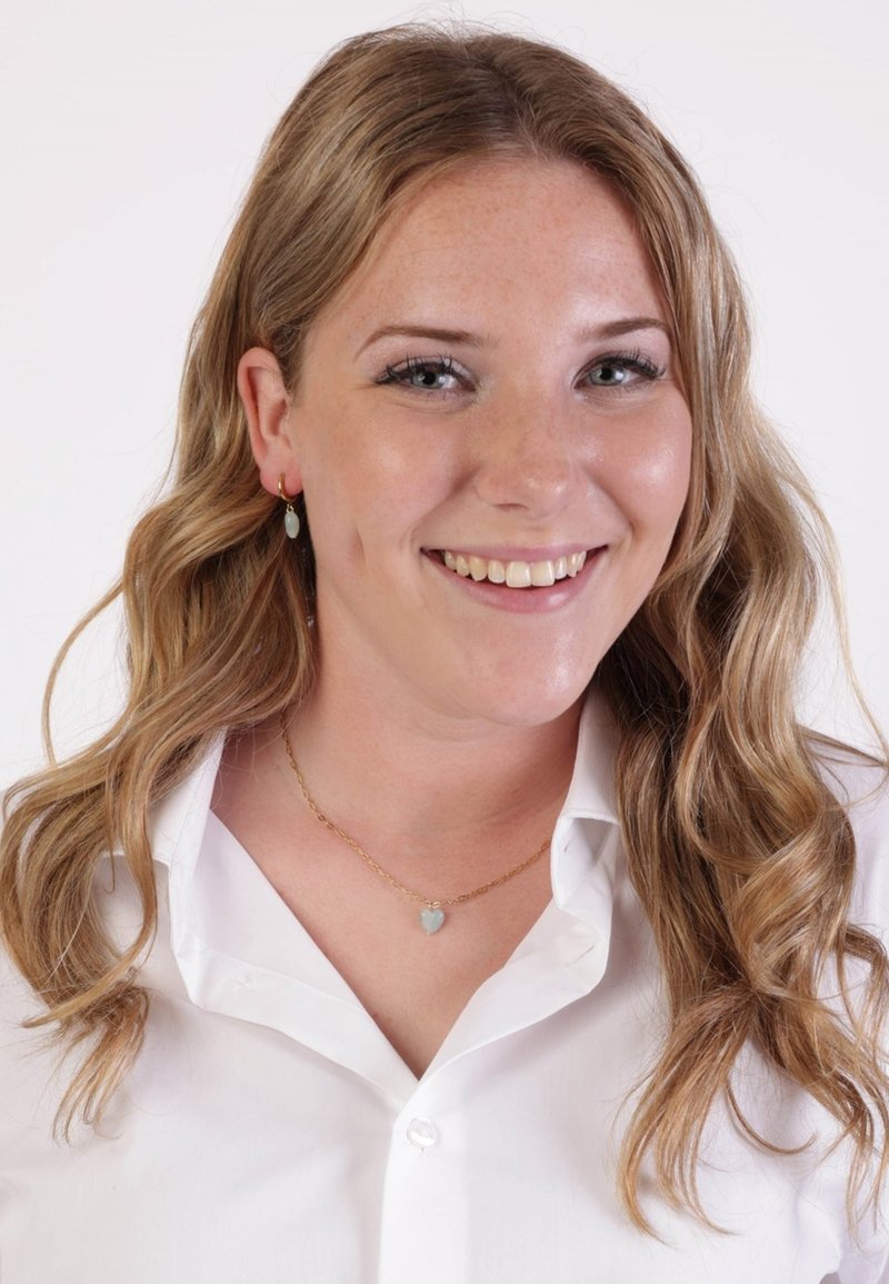Vrouw met golvend, blond haar, gekleed in een witte blouse. Ze heeft gouden oorbellen met lichtblauwe accenten en een bijpassende hartvormige ketting.