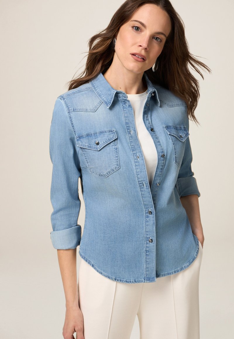 Camicia di jeans azzurro chiaro con colletto a punta, due tasche sul petto e maniche lunghe arrotolate, con bottoni metallici e orlo curvato.