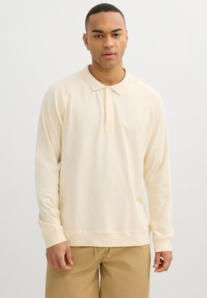 Joven con una camisa pólo crema de manga larga y pantalones marrón claro, de pie sobre un fondo blanco liso.