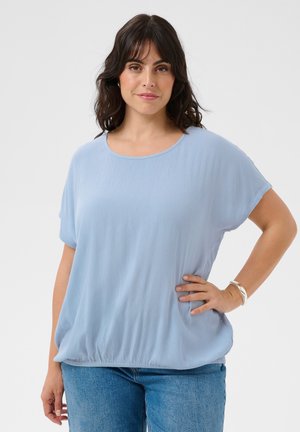 Frau mit dunklen Haaren, die eine lockere hellblaue Bluse und Jeans trägt, steht mit einer Hand in der Hüfte vor einem schlichten Hintergrund.