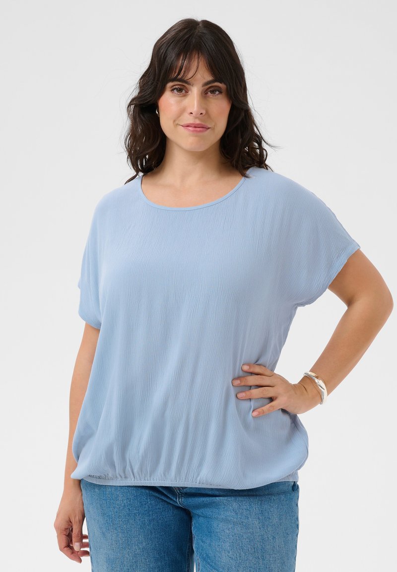 Femme aux cheveux foncés portant un chemisier bleu clair ample et un jean, debout avec une main sur la hanche sur un fond uni.