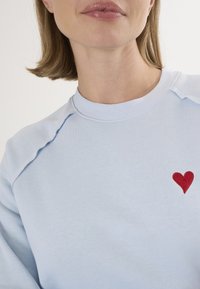 Felpa blu chiaro con collo rotondo a costine, caratterizzata da un cuore rosso ricamato sulla parte sinistra del petto e una texture morbida.