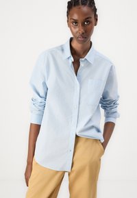 Camicia azzurra chiaro con bottoni e tasca, realizzata in un tessuto morbido. Presenta maniche arrotolate e una vestibilità ampia. Abbinata a pantaloni color khaki.