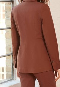 Blazer marron ajusté avec un design près du corps, des revers crantés et des accents de boutons sur les manches. Texture de tissu lisse.
