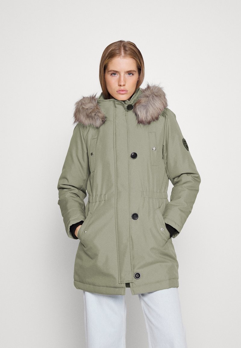 Femme portant un manteau d'hiver vert sauge avec une capuche à fourrure, un devant avec boutons et fermeture éclair, les mains dans les poches, et un pantalon bleu clair.