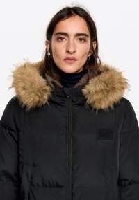 Czarna kurtka puchowa z odpinanym kapturem wykończonym beżowym futerkiem, zapięciem na zamek oraz naszywką z logo; tkanina o fakturze z pikowanymi detalami izolacyjnymi.