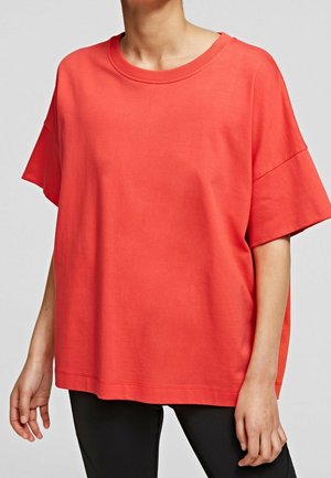 Henkilö pukeutuneena väljästi istuvaan, lyhythihaiseen korallinpunaisen väriseen T-paitaan mustien housujen päällä, yksiväristä harmaata taustaa vasten.