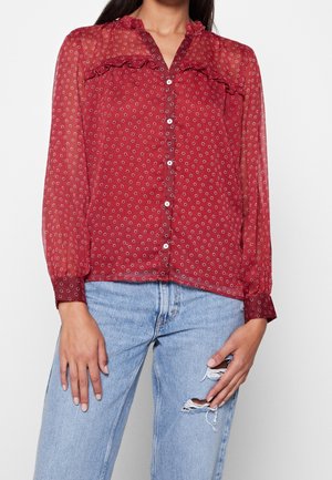 Kobieta w prześwitującej czerwonej bluzce z małymi kwiatowymi wzorami, falbankami, białymi guzikami i jasnoniebieskich podartych dżinsach.