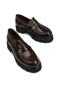 Brązowe skórzane mokasyny z okrągłym noskiem i ozdobnym paskiem. Grube czarne gumowe podeszwy z subtelnym wzorem bieżnika. Gładka faktura na całej powierzchni.
