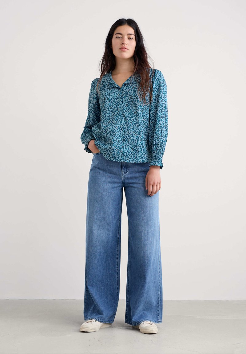 Sininen pitkähihaisen kukallisen kauluspaidan yhdistelmä leveälahkeisten vaaleansinisten farkkujen ja valkoisten tennareiden kanssa vaalealla taustalla.