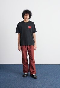 Sort bomulds t-shirt med rød grafik, parret med løse, skinnende, burgunder bukser. Modellen har sorte sneakers på. Enkel baggrund.