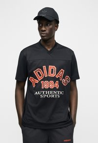 Άνδρας που φοράει μαύρο καπέλο Adidas, μαύρο διαπνέον μπλουζάκι με το κείμενο "ADIDAS 1994 AUTHENTIC SPORTS" και μαύρο σορτς με μικρό πορτοκαλί λογότυπο Adidas.