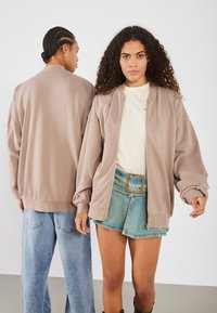 Light brown bomber jacket with a zip front, ribbed cuffs, and relaxed fit, worn over a cream shirt and paired with denim shorts.