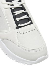 Zapatilla blanca con parte superior de cuero texturizado, acentos negros en el área de los cordones, lengüeta acolchada, suela de goma blanca y detalles de costura en contraste.