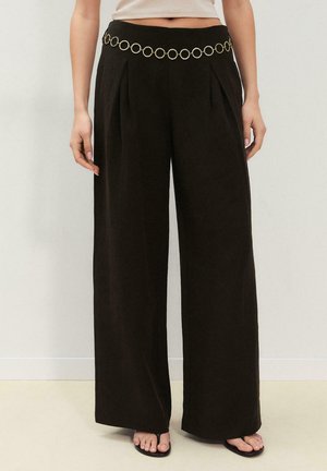 Kobieta w wysokich, czarnych spodniach z szerokimi nogawkami, posiadających przednie zakładki i złoty pasek łańcuszkowy, zestawionych z czarnymi sandałami na paski.