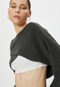 Maglione grigio a maglia testurizzata appoggiato su una canotta bianca, caratterizzato da una vestibilità ampia e un ampio scollo, con la pelle liscia visibile in vita.