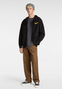 Schwarze Kapuzenjacke mit gelbem "VANS"-Logo, graues T-Shirt darunter, braune Hose, graue Beanie und schwarze Sneaker mit weißen Sohlen.