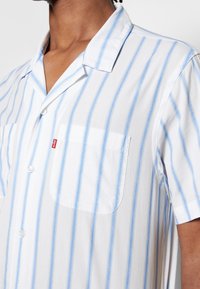 Camisa de manga corta con botones en blanco con rayas azul claro. Cuenta con un bolsillo en el pecho y cuello; fabricada con un tejido ligero.