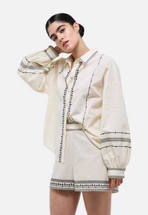 Jeune femme aux cheveux foncés en chignon bas porte une chemise crème oversize et un short avec des motifs brodés noirs et des boutons.