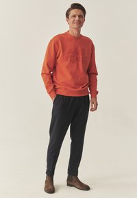 Orange sweatshirt med præget tekst, kombineret med mørkeblå bukser og brune ankelstøvler, hvilket skaber et afslappet outfit.