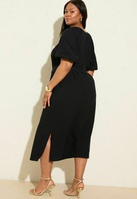 Schwarzes Midi-Kleid mit Puffärmeln, tailliertem Schnitt und seitlichen Schlitzen. Getragen mit goldenen Riemchensandalen und einem goldenen Armband am Handgelenk.