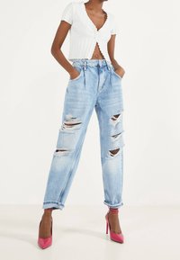 Helle blaue, ausgewaschene Jeans mit großen Rissen, im High-Waist-Design und umgeschlagenen Säumen. Kombiniert mit einem weißen, gerippten, kurz geschnittenen Oberteil und rosa High Heels.