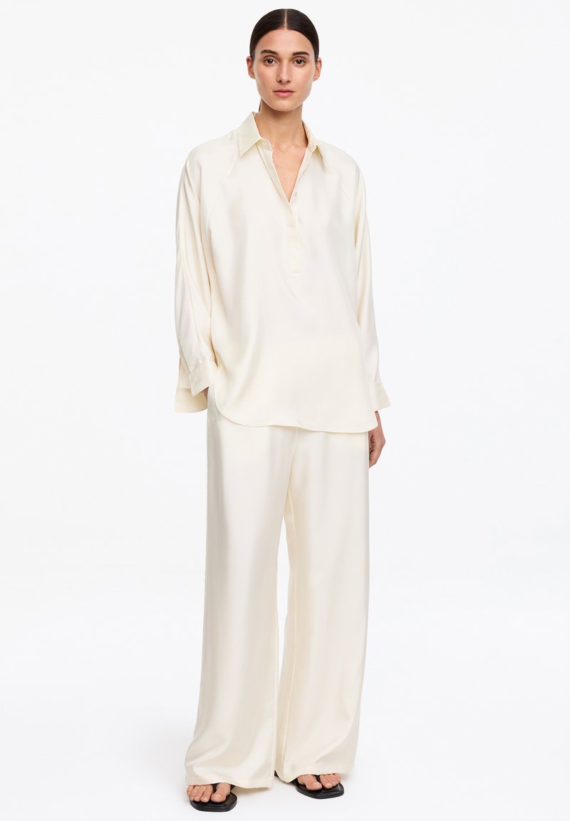 Femme aux cheveux foncés en chignon bas, portant une blouse ample en soie crème et un pantalon large assorti avec des sandales noires, debout sur fond blanc.