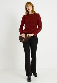 Pull en dentelle rouge à manches longues, coupe ajustée, associé à un pantalon noir évasé et à un petit sac à main en cuir de serpent marron avec une boucle dorée.