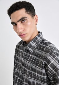Camicia a quadri nera e grigia con colletto strutturato, dotata di chiusura con bottoni sul davanti e un tessuto morbido e testurizzato.