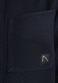 Donker marineblauwe textuurstof met een geweven patroon en een patchzak. Heeft een zwart label met een witte ontwerpaccent.