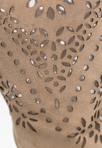 Beżowy zamszowy materiał z laserowo wyciętymi wzorami kwiatowymi, odsłaniającymi metaliczną warstwę pod spodem. Miękka tekstura z misternymi detalami wzoru.