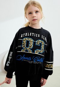 Svart sporttröja med blå och röda detaljer, stor leopardtryck "82" och texten "Athletics Club" på framsidan. Långa ärmar med detalj.