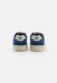 Egy pár sötétkék Adidas sporcipő fehér középtalppal, hátulról nézve, látható az Adidas logó a saroktabokon egy sima háttér előtt.