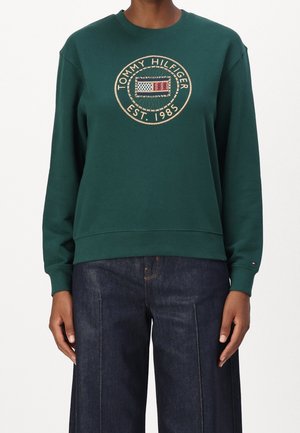 Osoba nosí zelenou mikinu s dlouhým rukávem s kulatým logem Tommy Hilfiger a tmavě modré džíny s širokými nohavicemi.