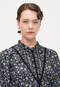 Blusa nera a fiori con colletto arricciato, caratterizzata da motivi floreali blu e verdi. Tessuto testurizzato con dettagli intrecciati lungo il collo.