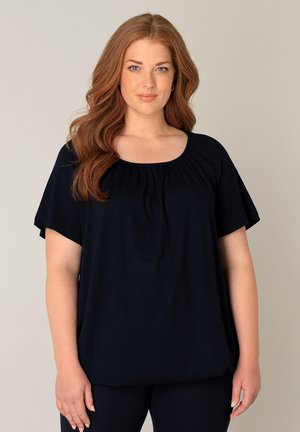 Vrouw met lang golvend rood haar, gekleed in een losse zwarte blouse met korte mouwen en een zwarte broek, staand tegen een effen beige achtergrond.