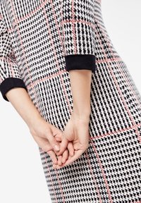 Robe pied-de-poule noir et blanc avec motif à carreaux rouges. Tissu en maille, manches mi-longues, poignets noirs, mains jointes à la taille.