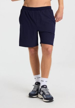 Underdel af en person iført marineblå atletikshorts, hvide sokker med tekst og blågrå løbesko på en hvid baggrund.