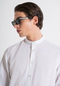 Camicia bianca in lino con colletto alla coreana e chiusura con bottoni, abbinata a occhiali da sole neri angolari con audaci accenti laterali.