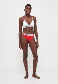 Fehér bikini felső "MOSCHINO I ❤️ YOU" felirattal, piros bikini alsóval párosítva. Mindkettő kontrasztos elasztikus derékpántokkal rendelkezik. Sima szövettextúra.